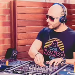 A man wearing sunglasses, headphones, and a "Café Mambo Ibiza" T-shirt, operating a Pioneer DJ mixer outdoors against a wooden slatted wall.