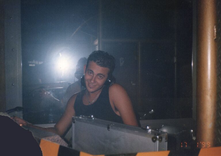 A flash photograph of a smiling young man wearing a black tank top and headphones, standing behind a DJ booth in a dark, hazy club environment.