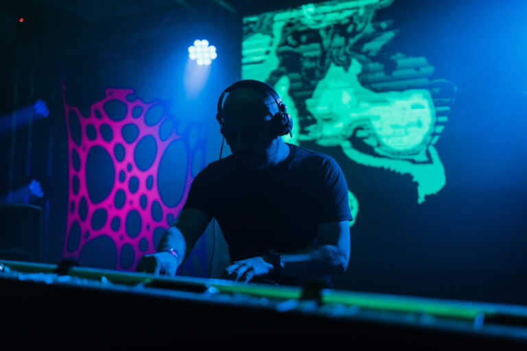A DJ wearing headphones performs behind a console in a darkly lit venue with vibrant blue and green stage lighting and abstract patterns on the background screens.