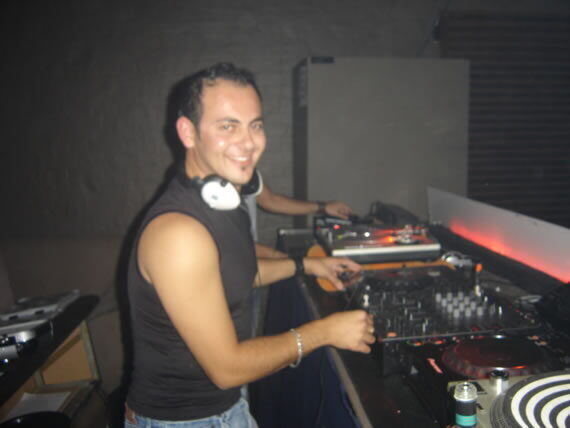 A smiling DJ in a black tank top and white headphones stands behind a professional mixing deck and turntables in a dimly lit venue.