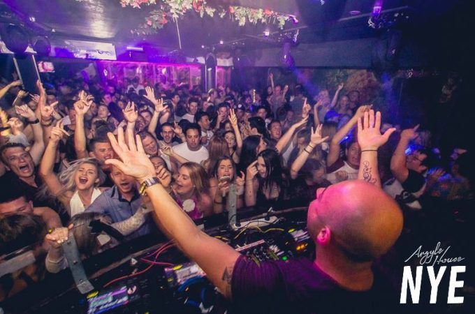 An over-the-shoulder view of a DJ with arms outstretched, facing a packed and enthusiastic dance floor.