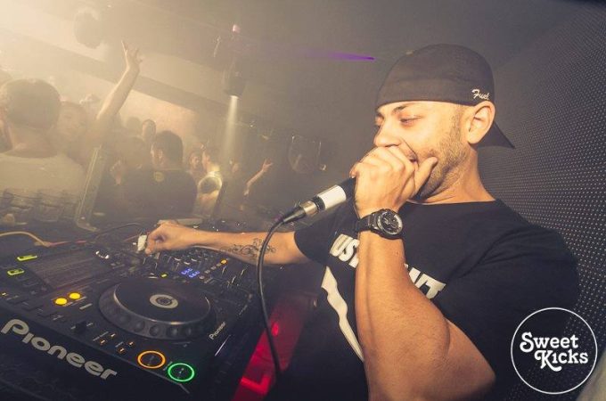 A DJ performing at a nightclub, wearing a black cap and t-shirt, speaking into a microphone while operating a Pioneer DJ mixer.