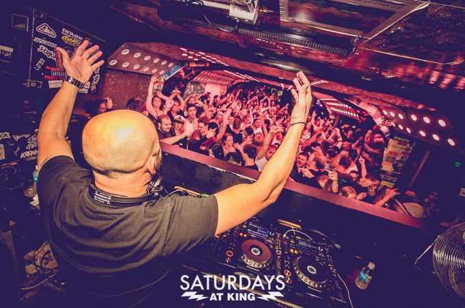 A DJ seen from behind with arms raised, performs for a crowded dance floor from a DJ booth during "SATURDAYS AT KING."