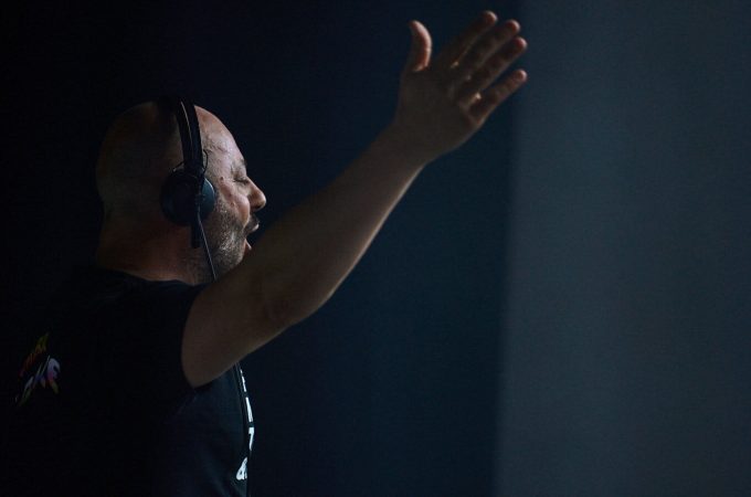 A side profile of DJ Fuel wearing headphones, with his arm raised and hand open, standing behind a soundboard in a dark setting.