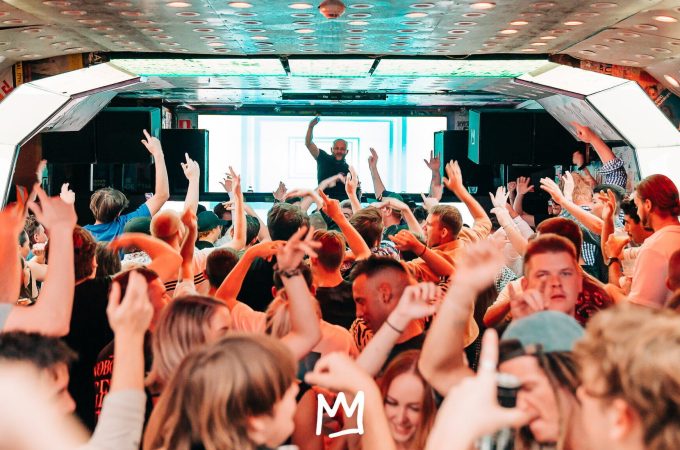 A high-energy crowd with arms raised dances in a futuristic, brightly lit club while a DJ performs on a stage in the background.