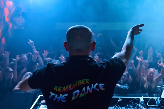 DJ wearing a "REMEMBER THE DANCE" shirt, looking out over a crowd with their hands in the air.