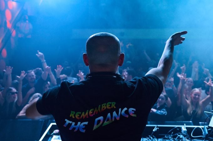 DJ wearing a "REMEMBER THE DANCE" shirt, looking out over a crowd with their hands in the air.