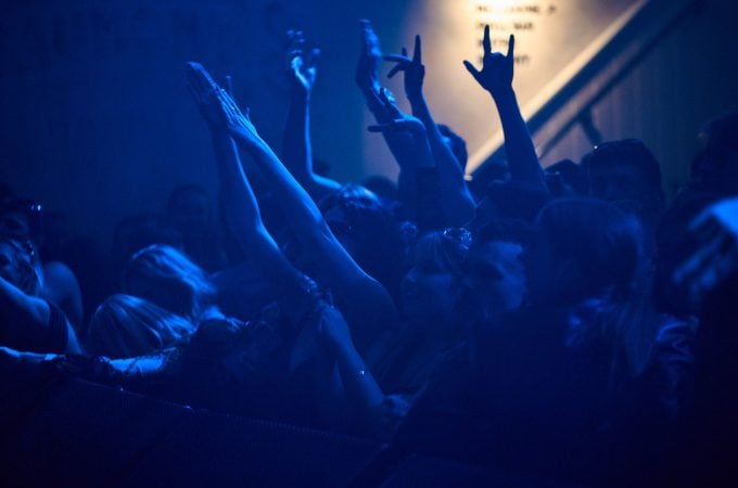 A crowd of people at a concert bathed in blue light, with many arms and hands raised in the air.