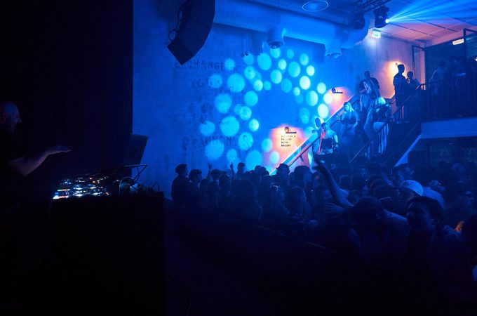 A DJ stands at a console in a dark club, performing for a large crowd illuminated by vibrant blue spotlights and a hazy atmosphere.