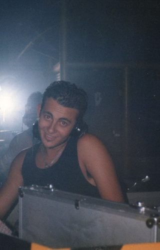 A flash photograph of a smiling young man wearing a black tank top and headphones, standing behind a DJ booth in a dark, hazy club environment.
