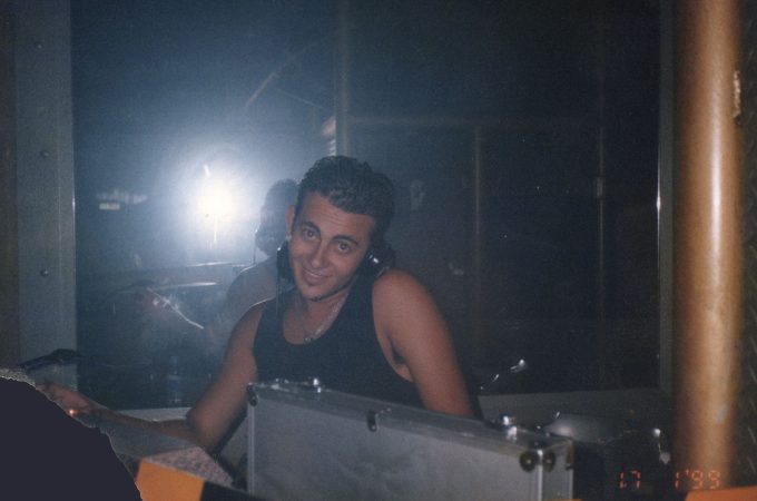 A flash photograph of a smiling young man wearing a black tank top and headphones, standing behind a DJ booth in a dark, hazy club environment.