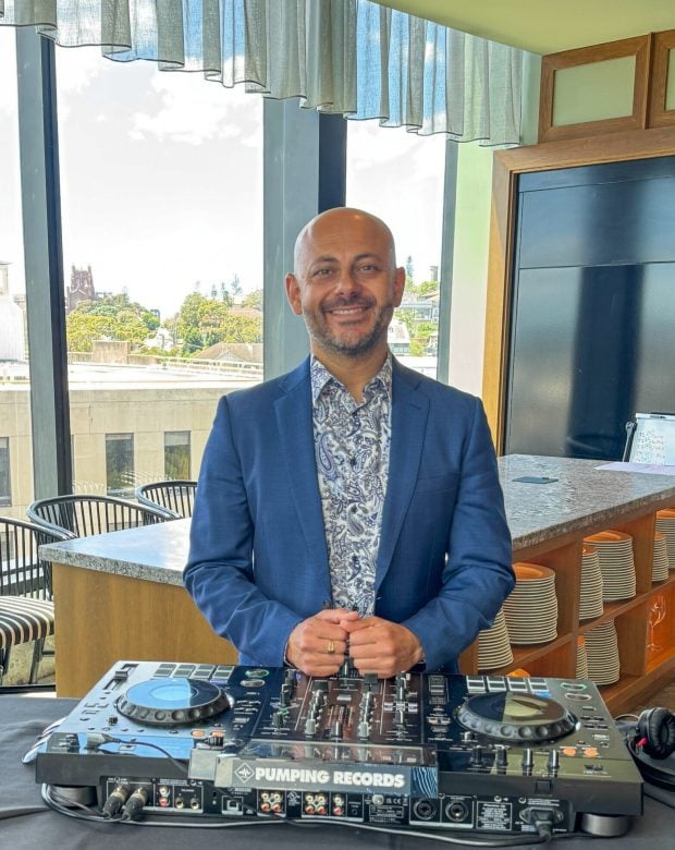 A smiling man in a blue blazer and paisley-patterned shirt stands behind a Pumping Records mixing deck in a bright room with large windows.