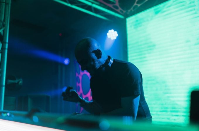 A side profile of a DJ wearing headphones around his neck, standing behind a lit-up deck against a backdrop of glowing green screens and blue stage lights.