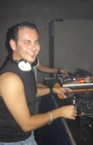 A smiling DJ in a black tank top and white headphones stands behind a professional mixing deck and turntables in a dimly lit venue.